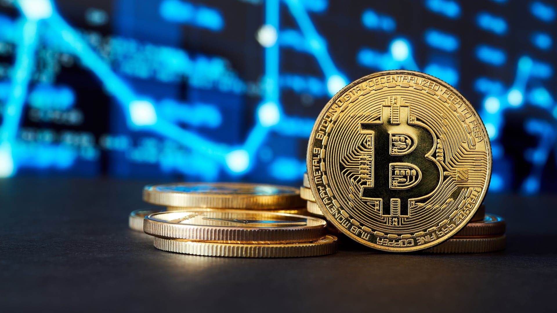 A gold Bitcoin coin standing beside a stack of coins, with a blue market chart behind it.